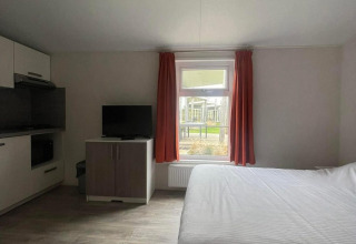 Lichte kamer in Comfort Lodge met tweepersoonsbed, televisie, raam met rode gordijnen en kitchenette.