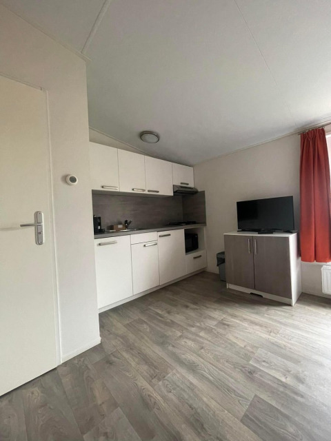 Stijlvol Comfort Lodge interieur met houten vloer, witte keuken, TV-meubel en rood gordijn. Honden welkom.