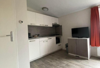Stijlvol Comfort Lodge interieur met houten vloer, witte keuken, TV-meubel en rood gordijn. Honden welkom.