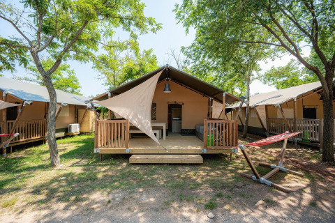 Safaritent met houten veranda en hangmat, gelegen tussen bomen op een zonnige glampingcamping.