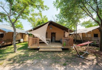 Safari-Zelt mit Holzveranda und Hängematte auf einem Campingplatz, umgeben von Bäumen und Sonne.