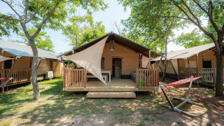 Tienda safari con porche de madera y hamaca, ubicada entre árboles en un camping glamping soleado.