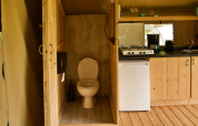 Interior view of a safari tent featuring a toilet, small kitchen area, gas stove, coffee maker, and mini fridge.