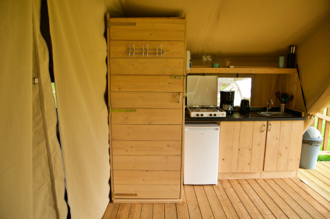 Interno di una tenda safari con angolo cottura in legno, fornello a gas, mini frigo e scaffale con stoviglie.