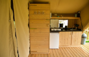 Interior of a safari tent featuring a wooden kitchenette, gas stove, mini fridge, and shelf with utensils.