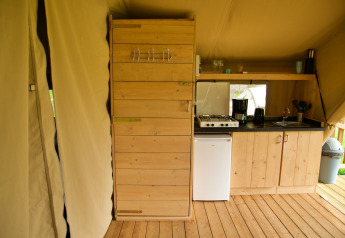Interno di una tenda safari con angolo cottura in legno, fornello a gas, mini frigo e scaffale con stoviglie.
