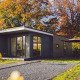 Moderne tiny house med store vinduer omgivet af træer i efterårsfarver og et udendørs siddeområde.