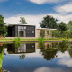 Moderne hytte ved sø i Vakantiepark Hölte, Holland, omgivet af natur og grønne planter under blå himmel.