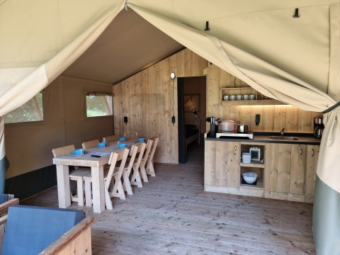 Binnenzicht van een glampingtent met houten eettafel, keuken, servies en zitruimte onder canvas dak.