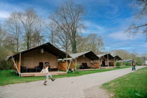 Een kind trapt een bal voor safaritenten met sanitair onder een mooi blauwe en licht bewolkte lucht.