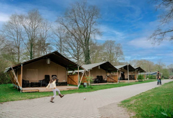 Een kind trapt een bal voor safaritenten met sanitair onder een mooi blauwe en licht bewolkte lucht.