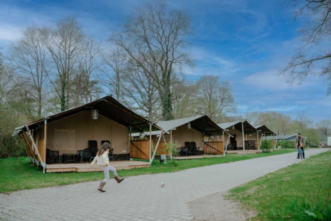 A child kicks a ball in front of safari tents with sanitary facilities under a blue sky with clouds.