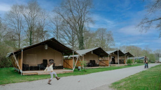 Un bambino gioca con una palla davanti a tende safari con servizi igienici sotto un cielo azzurro.