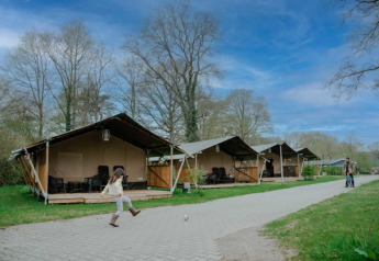 A child kicks a ball in front of safari tents with sanitary facilities under a blue sky with clouds.
