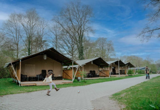 A child kicks a ball in front of safari tents with sanitary facilities under a blue sky with clouds.