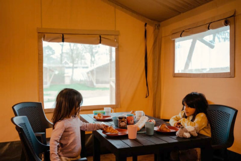 Twee kinderen eten ontbijt aan een tafel in een safaritent met ramen en zicht op buiten.