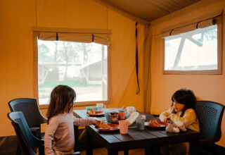 Twee kinderen eten ontbijt aan een tafel in een safaritent met ramen en zicht op buiten.