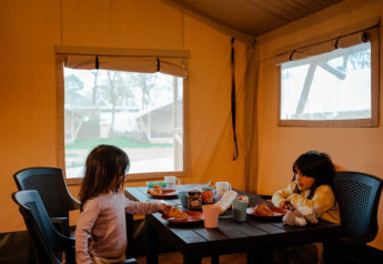 Two children have breakfast at a table inside a safari tent with windows and an outdoor view.