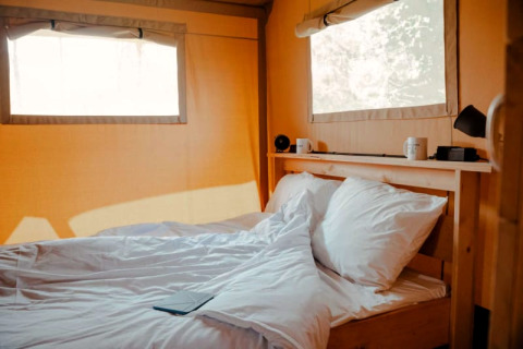 Binnenzicht van Safari tent + sanitary met gezellig bed, witte lakens en natuurlijke lichtinval.