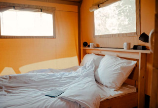 Binnenzicht van Safari tent + sanitary met gezellig bed, witte lakens en natuurlijke lichtinval.