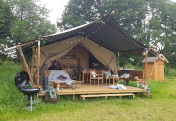 Tenda safari con veranda coperta, barbecue e zona relax immersi in una campagna verde e alberata.