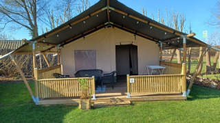 Tente safari avec véranda couverte, coin salon extérieur et clôture en bois sur pelouse verte.