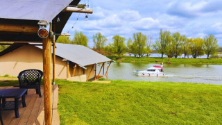 Tentes safari au bord d’un lac avec terrasse, vue sur un bateau et arbres verts en arrière-plan.