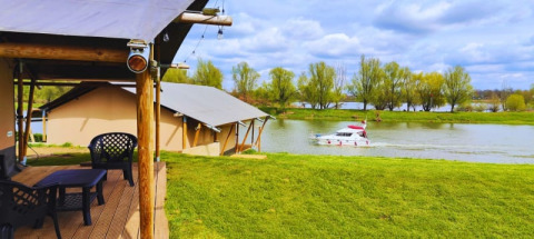 Tentes safari au bord d’un lac avec terrasse, vue sur un bateau et arbres verts en arrière-plan.