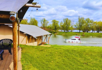 Safari tents by a lakeside with a deck, overlooking a boat on the water and lush green trees beyond.