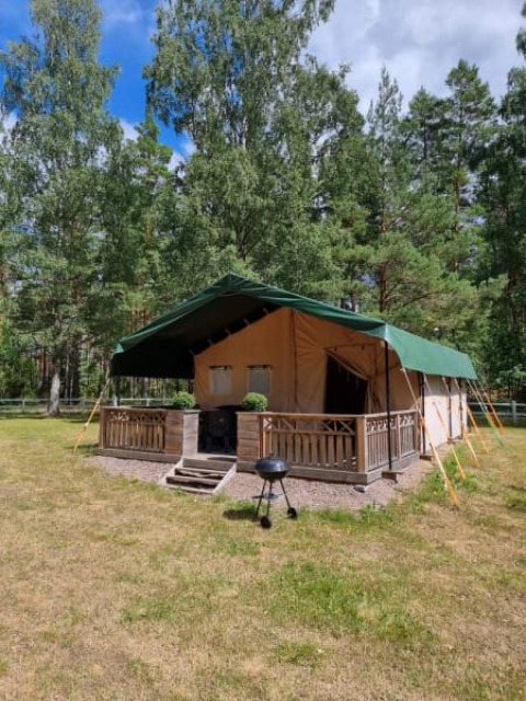 Safari-tent met sanitair, omgeven door groen bos, houten veranda en barbecue op het gazon vooraan.