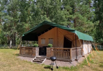 Tienda safari con instalaciones sanitarias en un entorno boscoso, porche de madera y parrilla exterior.