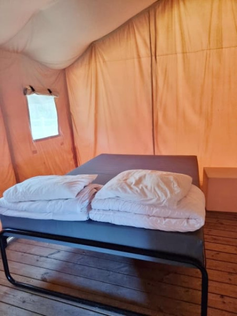Vista interior de una tienda safari mostrando una cama con sábanas y almohadas blancas sobre suelo de madera.