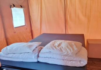 Vista interior de una tienda safari mostrando una cama con sábanas y almohadas blancas sobre suelo de madera.