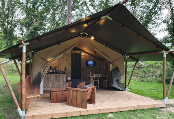 Tenda safari con servizi igienici, terrazza in legno e arredi immersa in un ambiente boscoso.