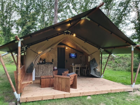 Safari tent with sanitary facilities, wooden deck and furniture set in a lush green forested area.