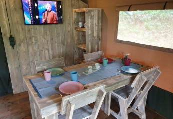 Dining area inside Safari tent with rustic wood table, dishes, TV and window at Camping het Horstmannsbos, Netherlands.