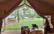 Uitzicht vanuit een safaritent met sanitair, houten terrasmeubilair en een groene camping buiten.