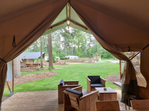 Blick aus einem Safari-Zelt mit Sanitäranlagen, Holzmöbeln auf der Terrasse und grünem Campingplatz.