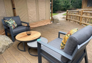 Zona de descanso exterior con sillones grises, mesa de madera y cojines estampados junto a tienda safari de lujo en Camping het Horstmannsbos.
