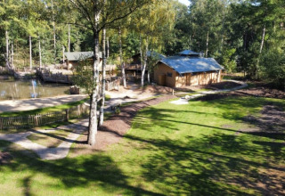 Safari tent area with grassy lawns, trees, a pond, and a wooden building in the background on a sunny day.