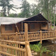 Safari tent with sanitary facilities on a wooden platform over a pond, surrounded by forest scenery.