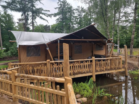 Tienda safari con instalaciones sanitarias, situada sobre una tarima de madera junto a un estanque y bosque.