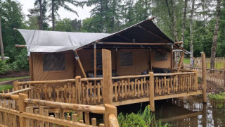 Safari tent with sanitary facilities on a wooden platform over a pond, surrounded by forest scenery.