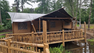Tienda safari con instalaciones sanitarias, situada sobre una tarima de madera junto a un estanque y bosque.