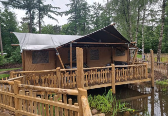 Tienda safari con instalaciones sanitarias, situada sobre una tarima de madera junto a un estanque y bosque.