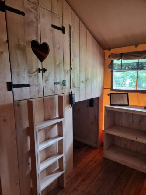 Interior de tienda safari con paredes de madera, estantería y escalera en Camping het Horstmannsbos, Países Bajos.