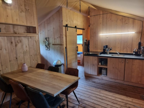 Vista interior de la tienda Safari en Camping het Horstmannsbos, con zona de comedor, cocina y suelos de madera.