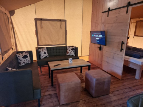 Sala en tienda safari con sofás, mesa y TV en Camping het Horstmannsbos, Países Bajos, ambiente acogedor.