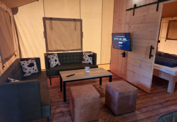 Sala en tienda safari con sofás, mesa y TV en Camping het Horstmannsbos, Países Bajos, ambiente acogedor.