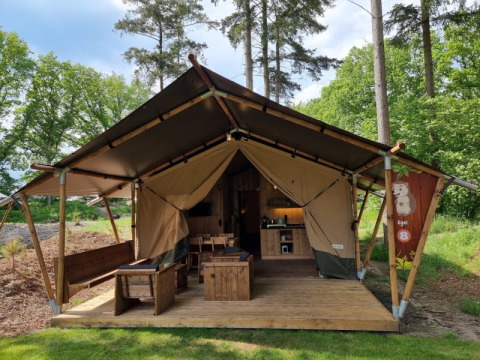 Tenda safari con servizi igienici, veranda in legno e arredi, situata in un campeggio tra gli alberi.